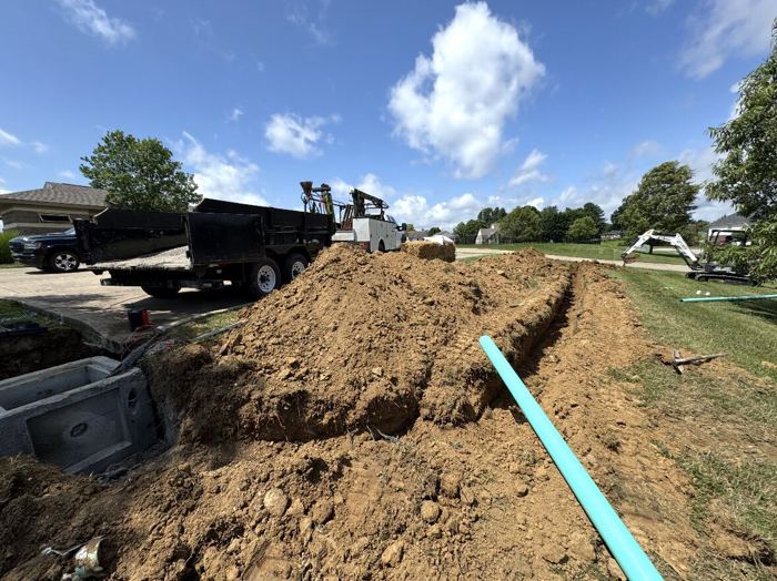Open trench across a front yard with new blue water service line laid in, a dump truck and mini excavator on the job site