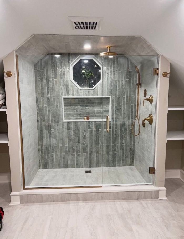 Finished walk-in tile shower with grey stone tile, brushed brass rain head and fixtures, built-in niche and frameless glass door